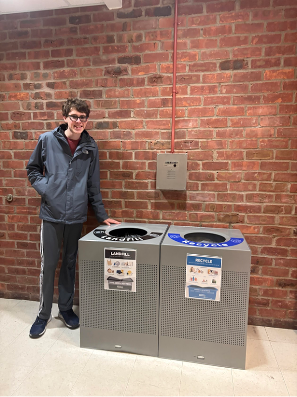 Student standing next to labeled landfill and recycling bins against a brick wall.