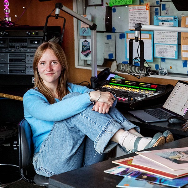 A student sitting cross-legged in a radio studio, smiling beside a microphone and soundboard.