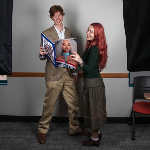 Two people stand in a studio setting, smiling and looking at a magazine together with a portrait on the cover.