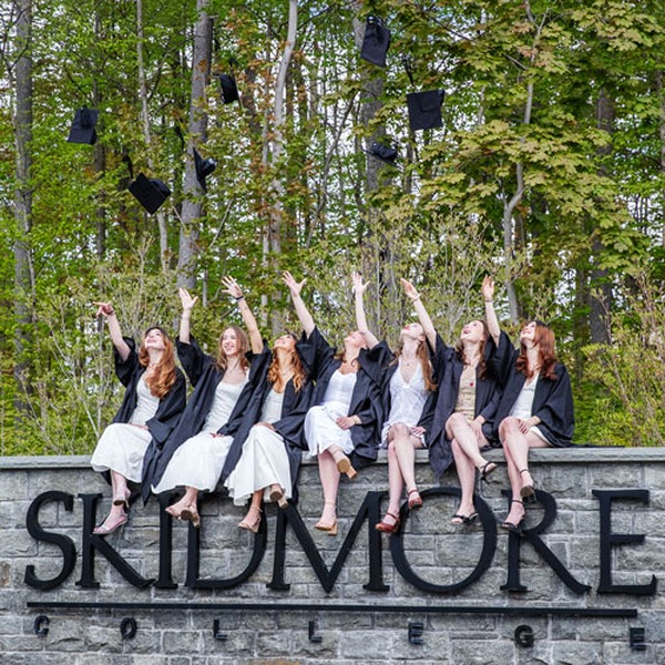 A group of graduates in caps and gowns celebrate by tossing their graduation caps into the air while sitting on a stone sign 