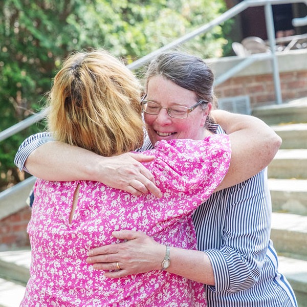 Two colleagues embrace warmly outside near a campus stairway, smiling as they hug during a celebration.