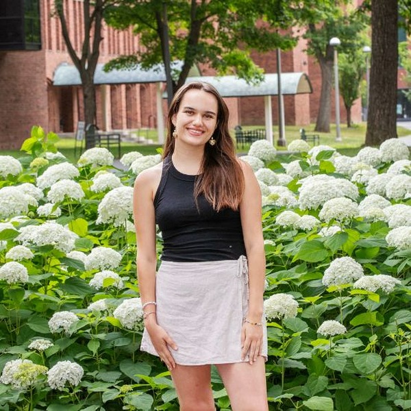 A young woman stands smiling in front of white hydrangeas. She is wearing a black sleeveless tank and a gray skirt.
