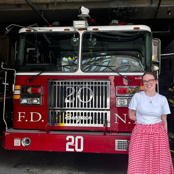 A smiling woman wearing glasses, a white shirt, and a red-and-white checkered skirt stands in front of a bright red fire truc