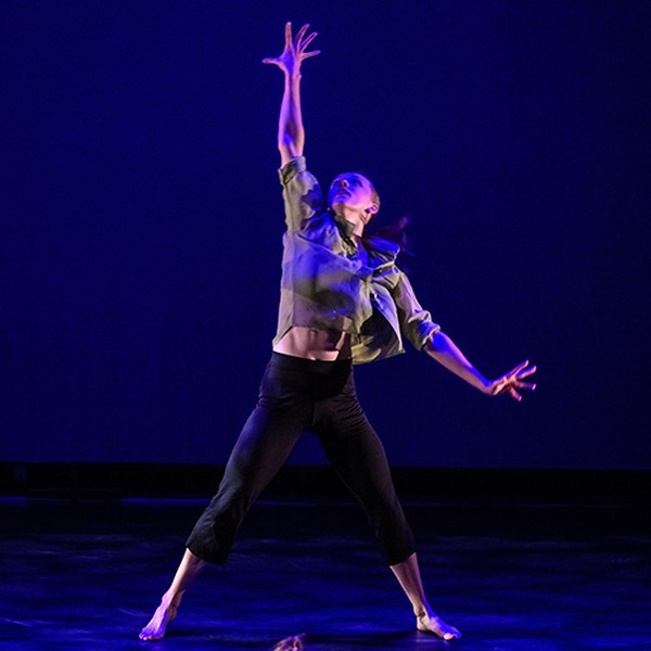 Barefoot contemporary dancer in black pants and a green shirt reaches one arm upward under blue stage lighting.
