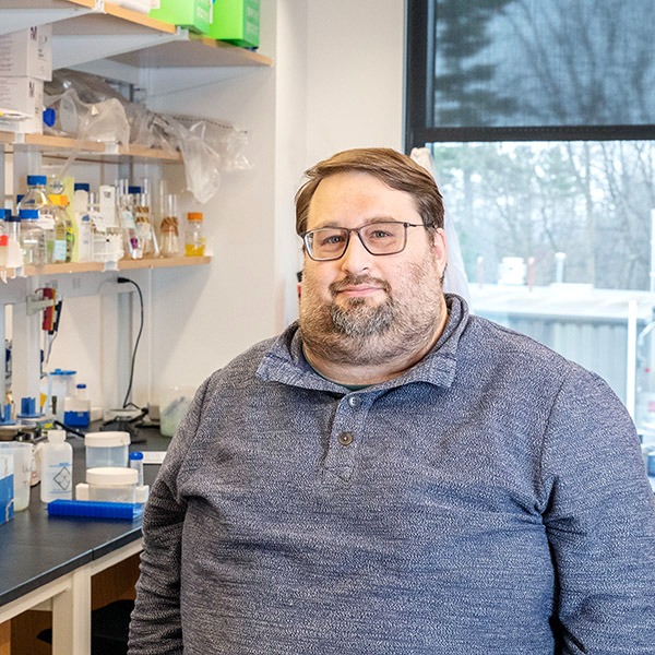 Person with glasses seated at a laboratory bench, with shelves of scientific equipment and bottles in the background.