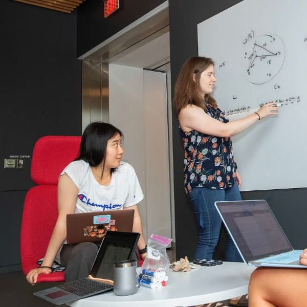 Kirsten Hogenson writes equations on a white board as Riley Vavolizza and Rachel Xia watch with computers on their laps