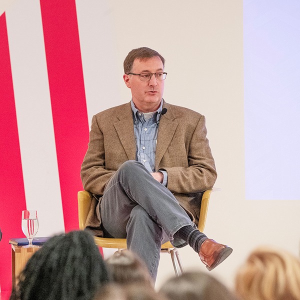 Christopher Mann seated with legs crossed in a chair on stage, speaking during a panel, wearing a tan blazer, blue shirt, and